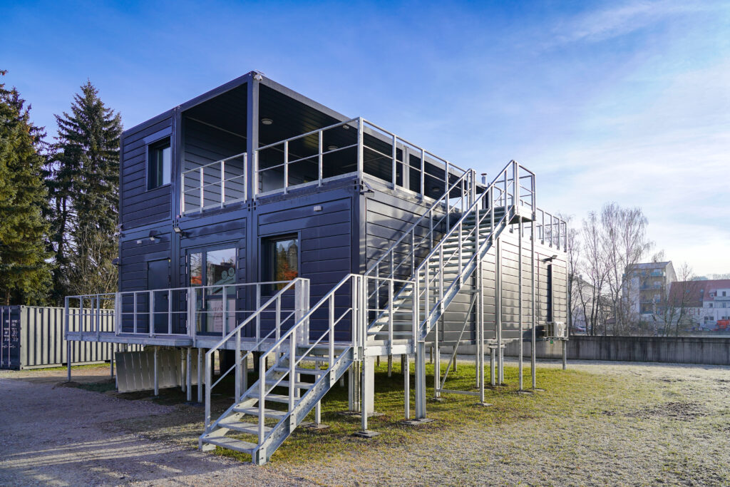 Ein modernes, zweistöckiges Gebäude aus gestapelten Schiffscontainern mit Metalltreppen und Geländern, das auf Stelzen steht, befindet sich an einem sonnigen Tag im Freien in der Nähe von Bäumen und anderen Gebäuden.