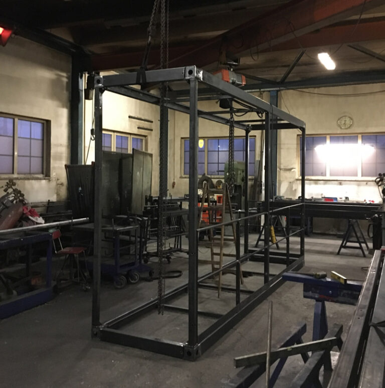 Ein großer rechteckiger Metallrahmen, der an Ketten im Inneren einer Werkstatt aufgehängt ist, in der Werkzeuge, Werkbänke und Leitern zu sehen sind. Der Raum ist schwach beleuchtet, hat mehrere Fenster und eine Uhr an der Wand.