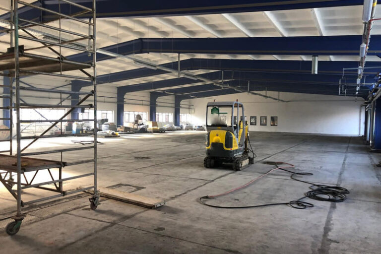 Ein großes, leeres Industrielager mit freiliegenden Balken, verstreuten Baumaschinen, einer gelben Bodenreinigungsmaschine und Gerüsten auf der linken Seite. Das Sonnenlicht fällt durch die Fenster an der Wand.