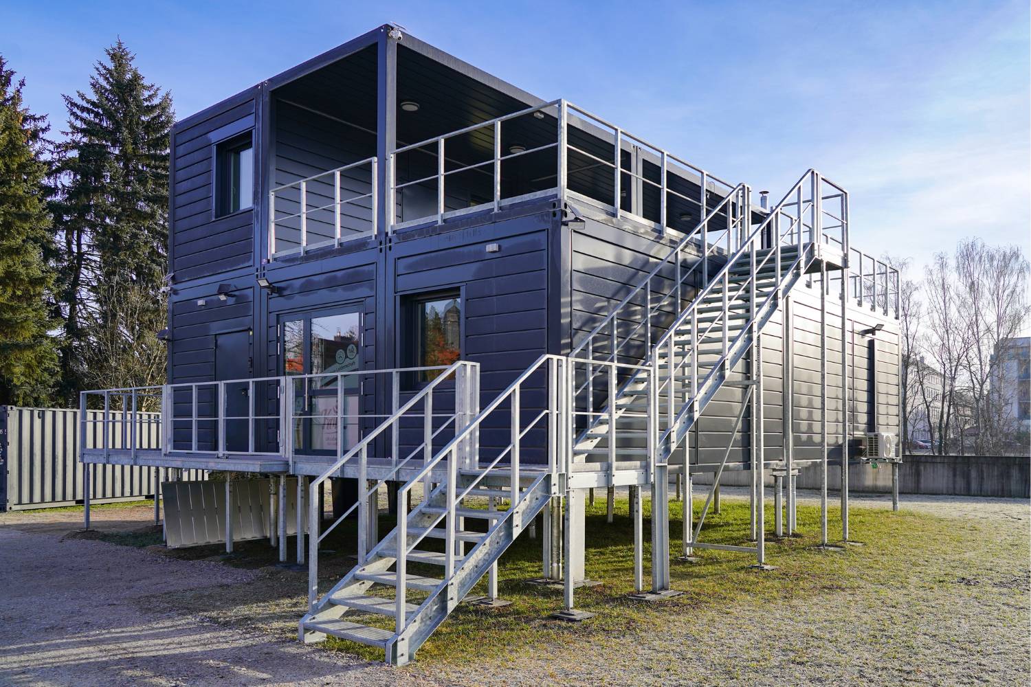 Ein modernes zweistöckiges modulares Gebäude mit Metallfassade, großen Fenstern und mehreren Außentreppen, das auf erhöhten Stützen auf einer Wiese mit Bäumen und freiem Himmel steht.