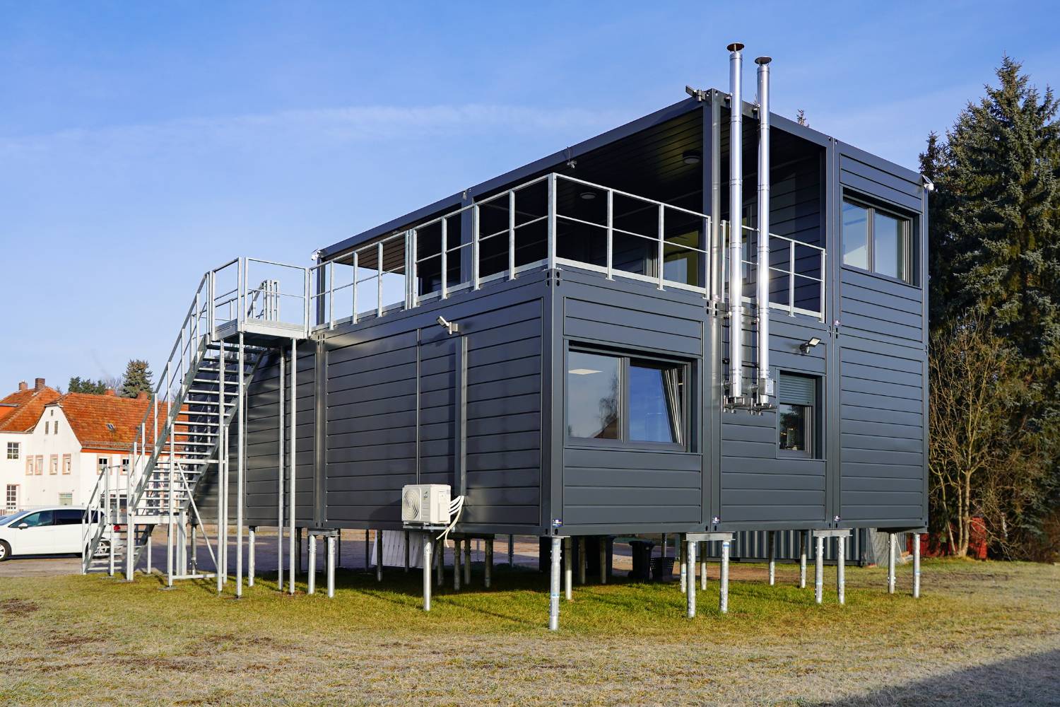 Ein modernes, graues, zweistöckiges Haus in Modulbauweise auf Metallstelzen mit Außentreppe, großen Fenstern und Metallgeländern, das auf einer Wiese mit Bäumen und anderen Gebäuden im Hintergrund unter freiem Himmel steht.