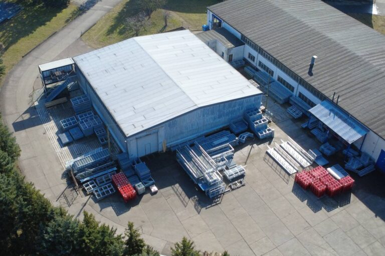 Luftaufnahme einer Industrieanlage mit gestapelten Metallrahmen und roten Containern im Freien, umgeben von Bäumen und gepflasterten Flächen, angrenzend an ein großes Lagergebäude.