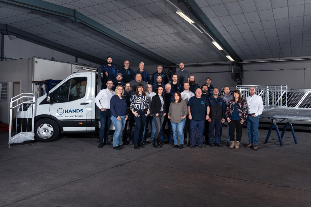 Eine Gruppe von etwa 25 Personen posiert für ein Foto in einem Industrielager, neben einem weißen Lieferwagen mit der Aufschrift HANDS und einem Metallgeländer stehend. Bei der Gruppe scheint es sich um Mitarbeiter des Unternehmens zu handeln.