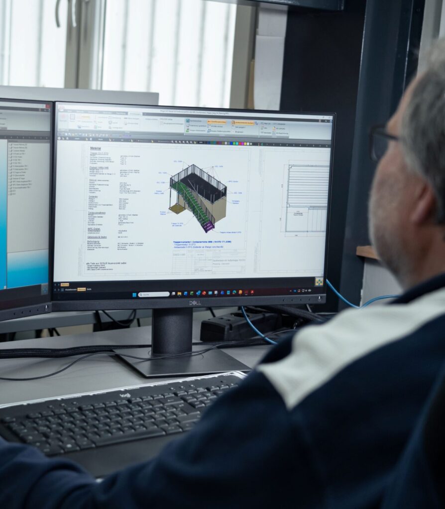 Eine Person, die an einem Schreibtisch sitzt, verwendet einen Computer mit einem großen Monitor, auf dem ein architektonischer oder technischer 3D-Entwurf mit technischen Details und Diagrammen angezeigt wird.