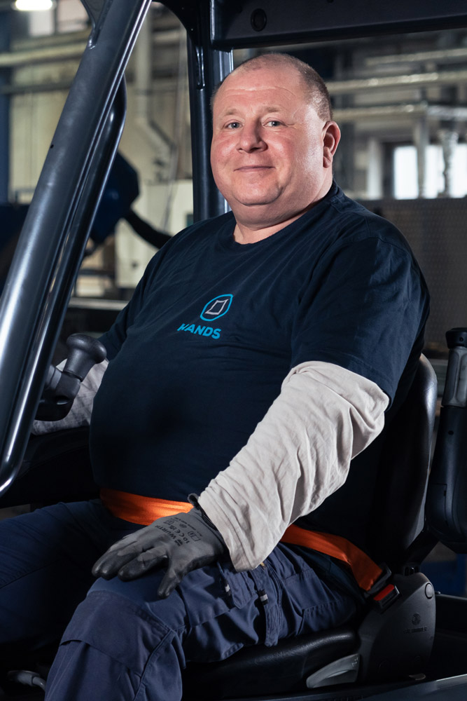 Ein lächelnder Mann mit dunklem T-Shirt und Arbeitshandschuhen sitzt auf einem Gabelstapler in einer Industrieumgebung und schaut in die Kamera.