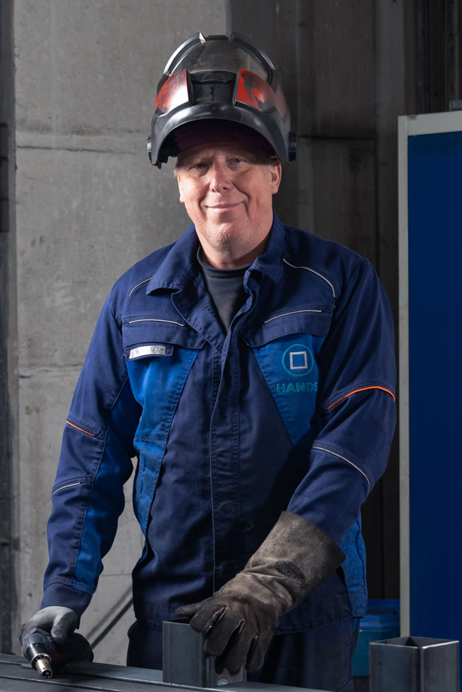 Ein lächelnder älterer Mann in einer blauen Arbeitsuniform und einem Schweißerhelm steht in einer Halle, trägt Handschuhe und stützt seine Hände auf eine Metallfläche.