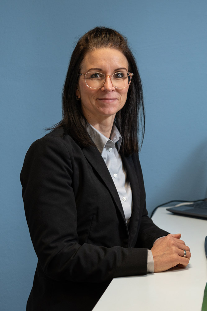 Eine Frau mit langen dunklen Haaren, die eine Brille, einen schwarzen Blazer und ein hellgraues Hemd trägt, sitzt an einem weißen Schreibtisch vor einer blauen Wand, schaut in die Kamera und lächelt leicht.