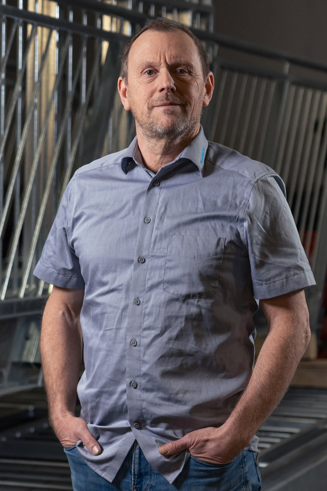Ein Mann mittleren Alters steht in einem Haus, hat die Hände in den Jeanstaschen und trägt ein hellgraues, kurzärmeliges Button-up-Hemd. Im Hintergrund sind Metallgeländer und industrielle Elemente zu sehen.