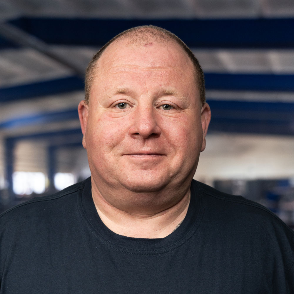 Ein Mann mittleren Alters mit kurzen Haaren und einem leichten Lächeln steht in einem Raum und trägt ein dunkles T-Shirt. Der Hintergrund ist unscharf und zeigt einen großen Raum mit blauen Balken und Fenstern.