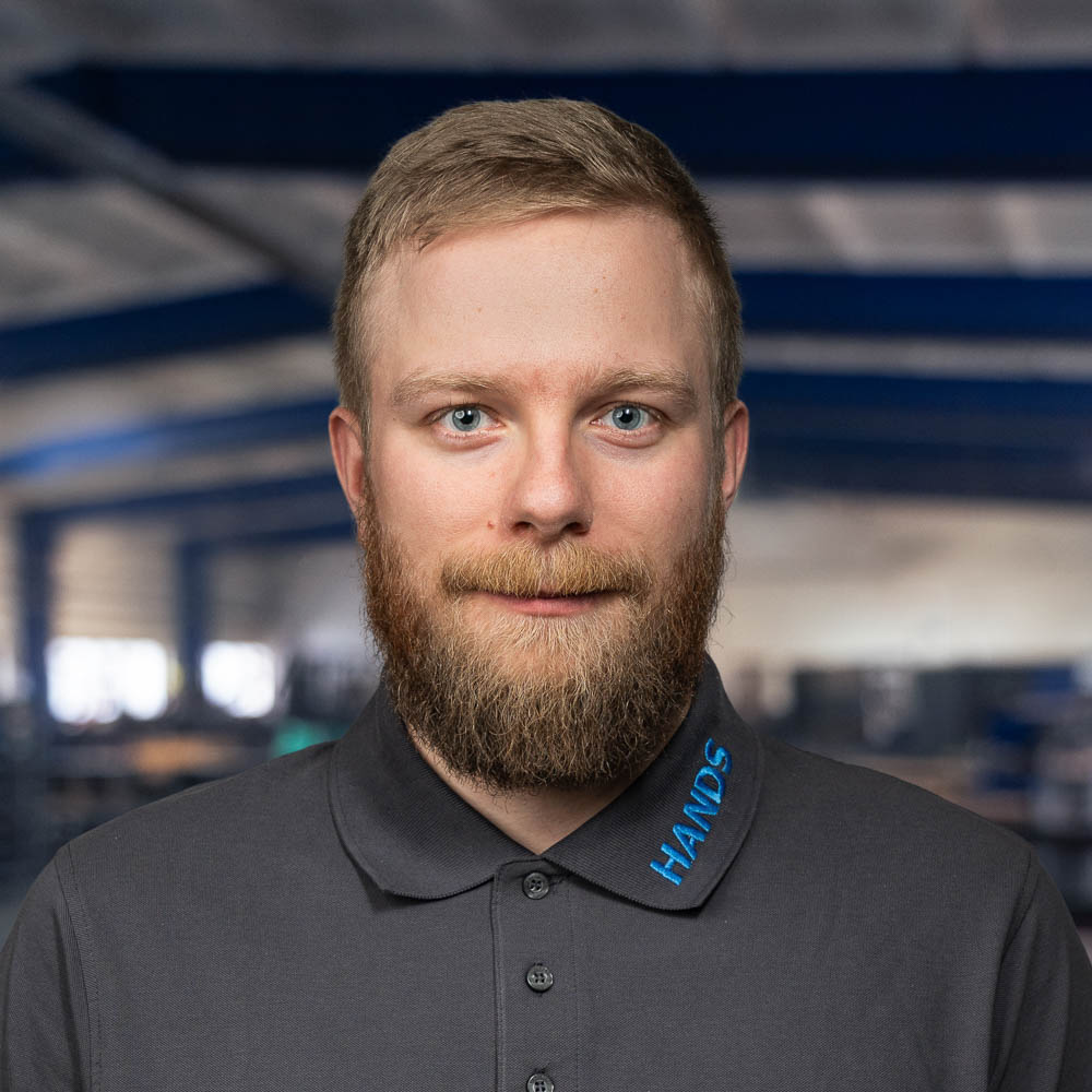 Ein bärtiger Mann mit kurzen blonden Haaren, der ein graues Hemd mit Kragen trägt, auf dessen Kragen „HANDS“ gestickt ist, steht in einem Innenraum, im Hintergrund sind verschwommene Balken im Industriestil zu sehen.
