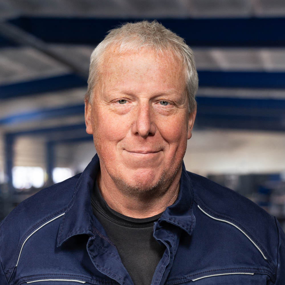 Ein Mann mittleren Alters mit kurzen grauen Haaren und einer blauen Jacke lächelt sanft, während er in einem großen, industriell anmutenden Gebäude mit blauer Stahlkonstruktion steht.