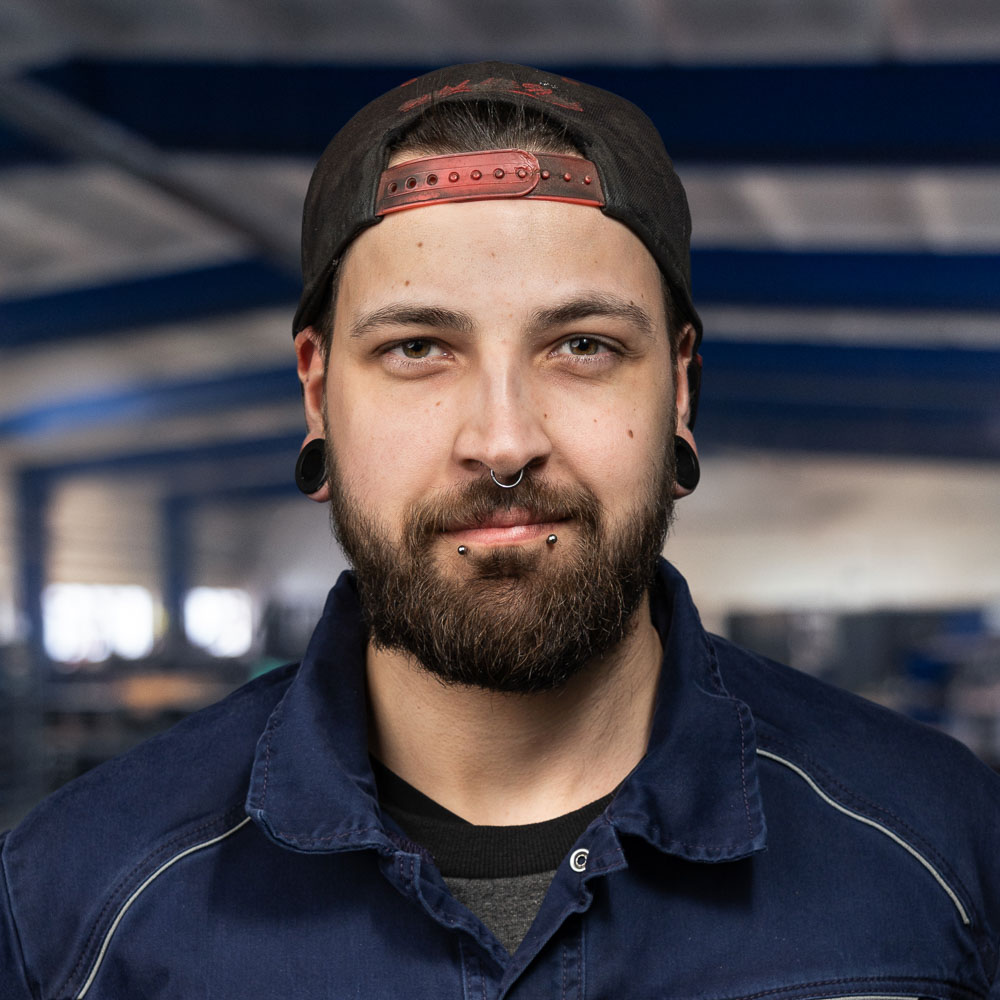 Ein Mann mit Bart, Piercings und gedehnten Ohrläppchen trägt eine dunkle Kappe verkehrt herum und eine marineblaue Arbeitsjacke; er steht in einem gut beleuchteten Industriegebäude.