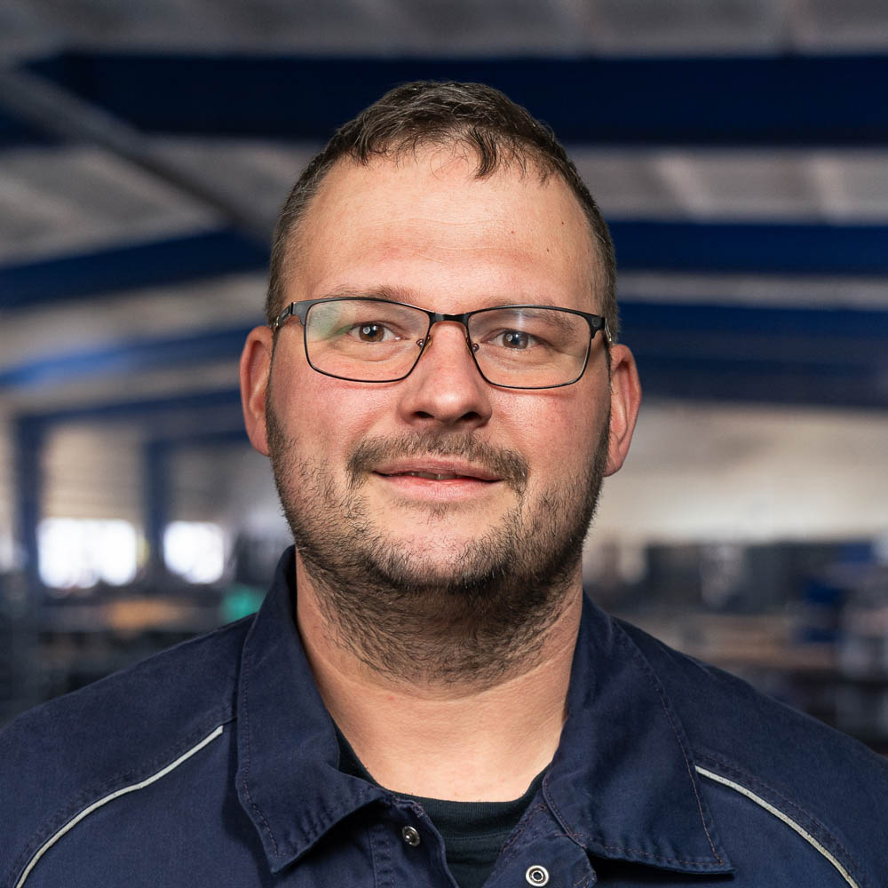 Ein Mann mit kurzen Haaren, Brille und einem gepflegten Bart trägt eine dunkle Arbeitsjacke und steht in einem Innenraum vor einem verschwommenen industriellen Hintergrund, über ihm sind blaue Lichtstrahlen zu sehen.