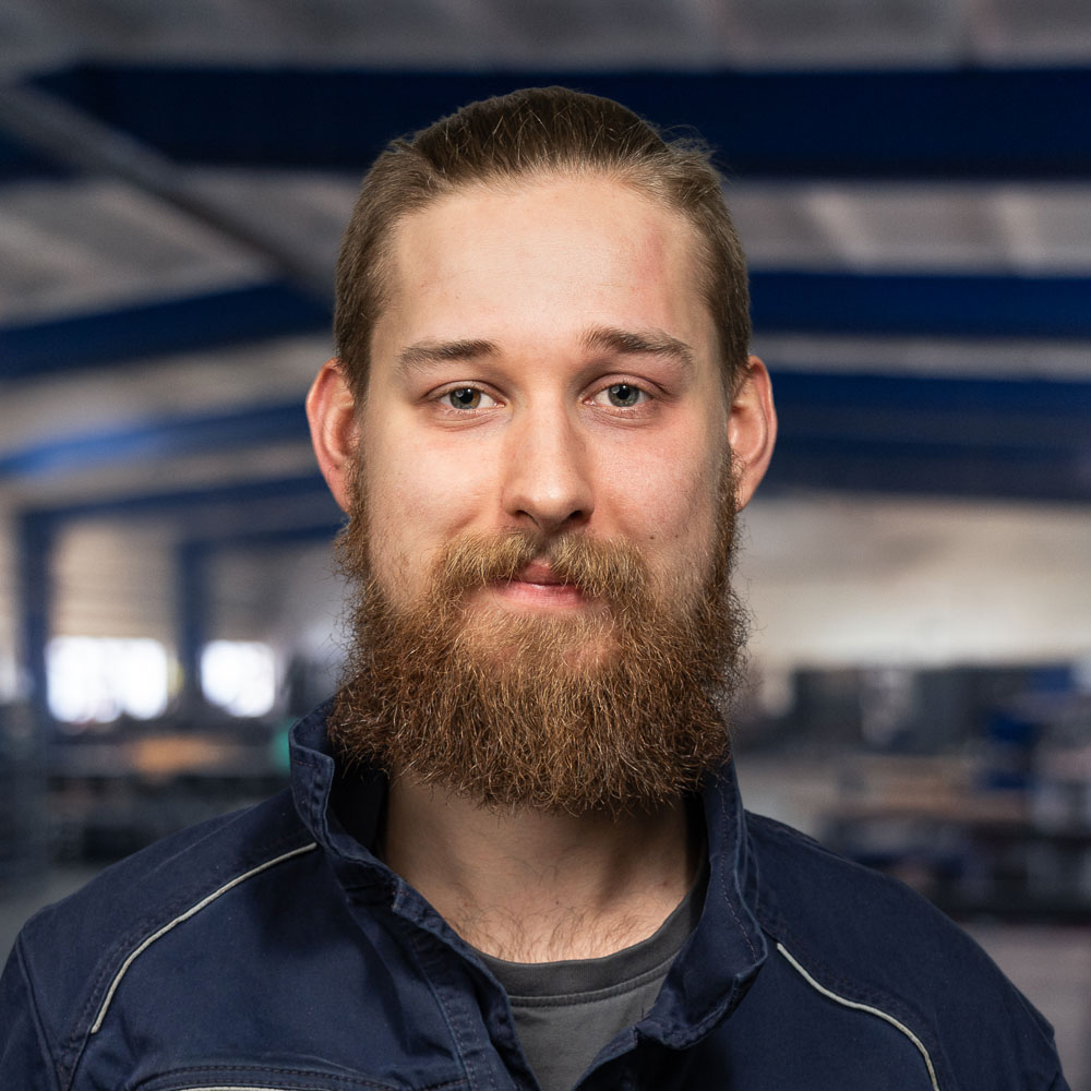Ein junger Mann mit braunem Bart und Schnurrbart, der eine dunkelblaue Arbeitsjacke trägt, steht in einer großen Industrie- oder Lagerhalle und lächelt leicht in die Kamera.