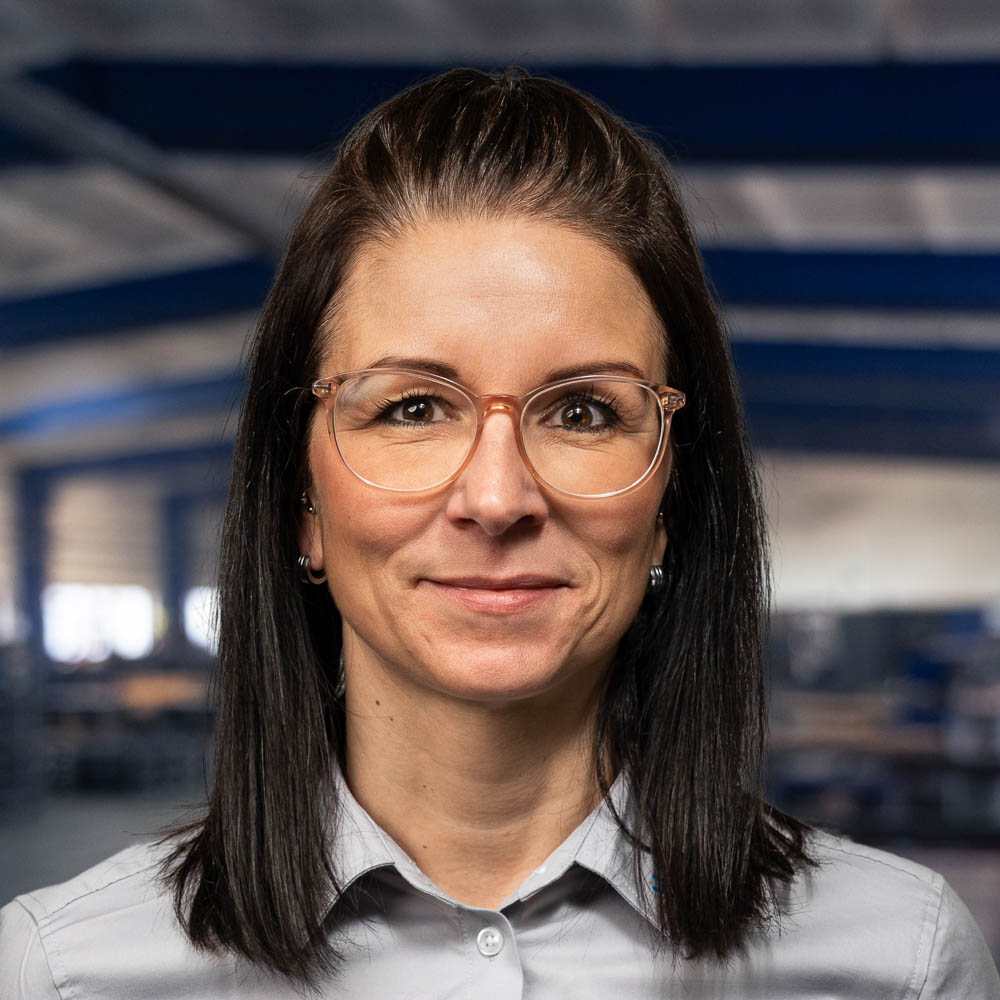 Eine Frau mit glattem, dunklem Haar und Brille lächelt leicht; sie trägt ein hellgraues Hemd mit Kragen. Sie befindet sich in einem Innenraum, im Hintergrund ist verschwommen eine Industrie- oder Lagerhalle zu erkennen.