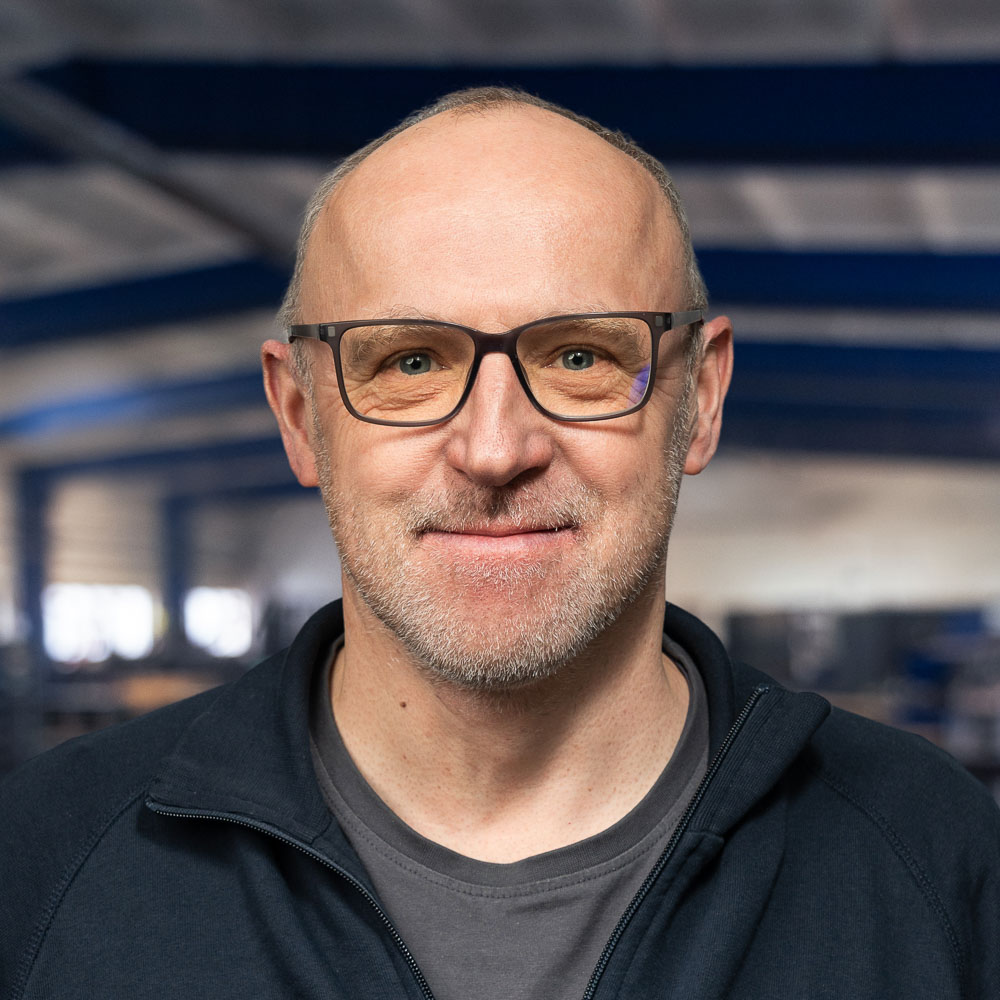 Ein kahlköpfiger Mann mit Brille, der eine dunkle Jacke und ein T-Shirt trägt und sanft lächelt, während er in einem großen, modernen Industriegebäude steht, im Hintergrund sind blaue Balken zu sehen.