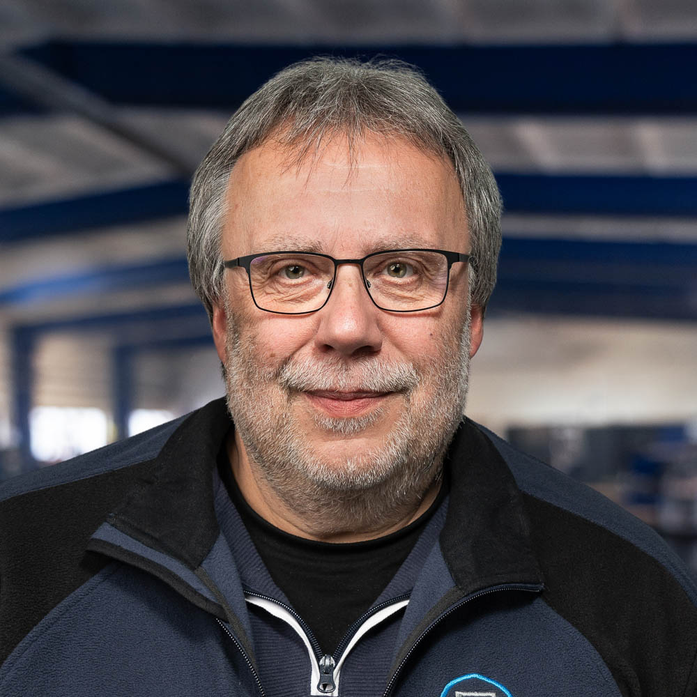 Ein Mann mittleren Alters mit grauen Haaren, Brille und Bart lächelt in einer blauen Industrieumgebung in die Kamera. Er trägt eine dunkle Jacke über einem marineblauen Hemd.