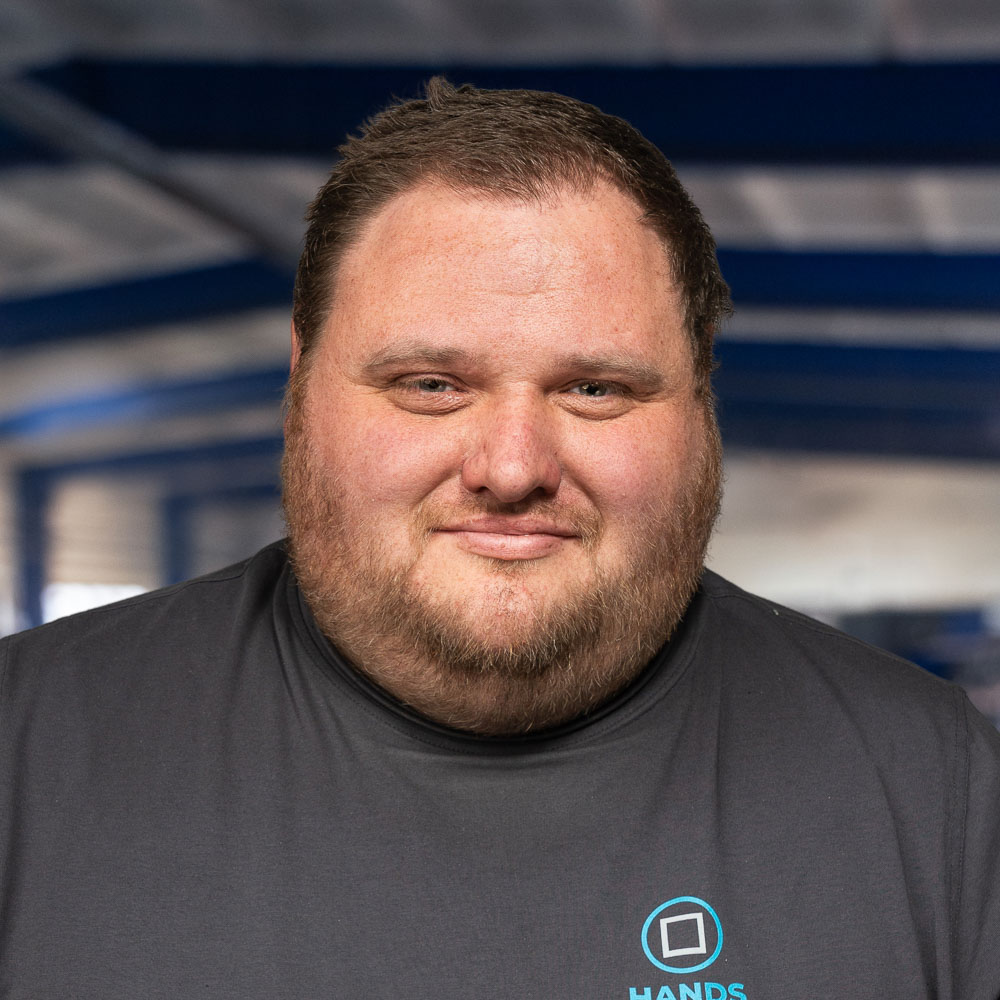 Ein Mann mit kurzen braunen Haaren und Bart lächelt sanft; er trägt ein dunkles T-Shirt und steht in einem Raum vor einem verschwommenen blau-weißen Hintergrund.