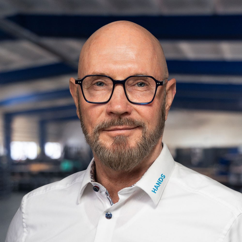 Ein kahlköpfiger Mann mit Brille und kurzem Bart, der ein weißes Hemd mit der Aufschrift „HANDS“ am Kragen trägt und in einer großen, unscharfen Industrie- oder Lagerhalle steht.