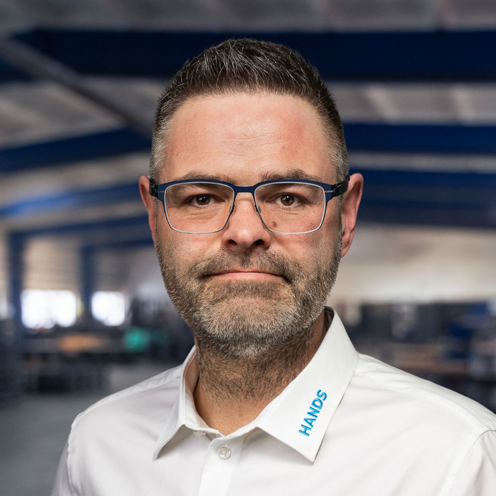 Ein Mann mit kurzen braunen Haaren, Brille und Bart, der ein weißes Hemd mit der Aufschrift „HANDS“ am Kragen trägt, steht in einer Industrieumgebung mit blauen Lichtstrahlen und unscharfem Hintergrund.