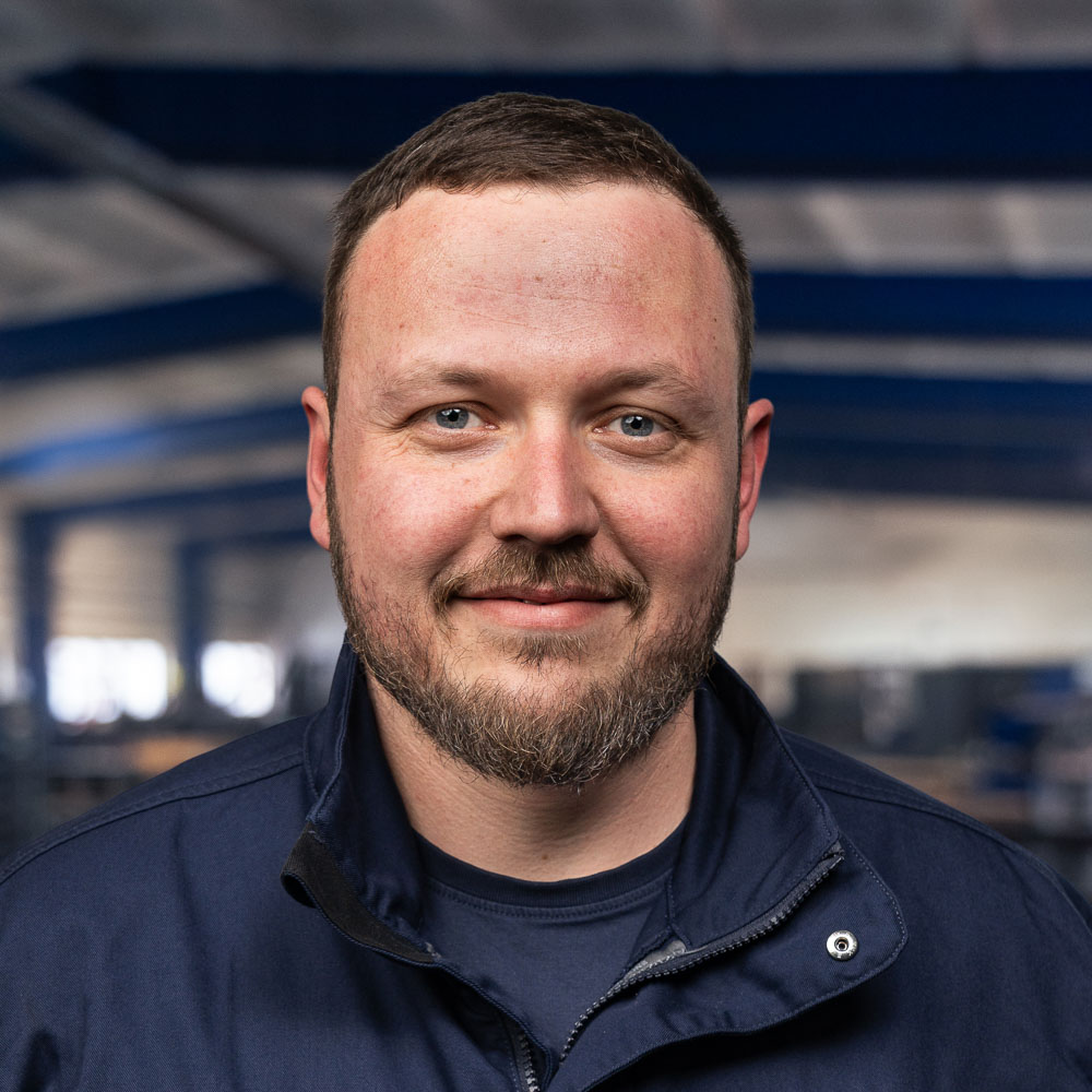 Ein Mann mit kurzen braunen Haaren und Bart, der eine marineblaue Jacke trägt, steht in einem geräumigen Industriegebäude mit blauen Trägern und sanfter Beleuchtung.
