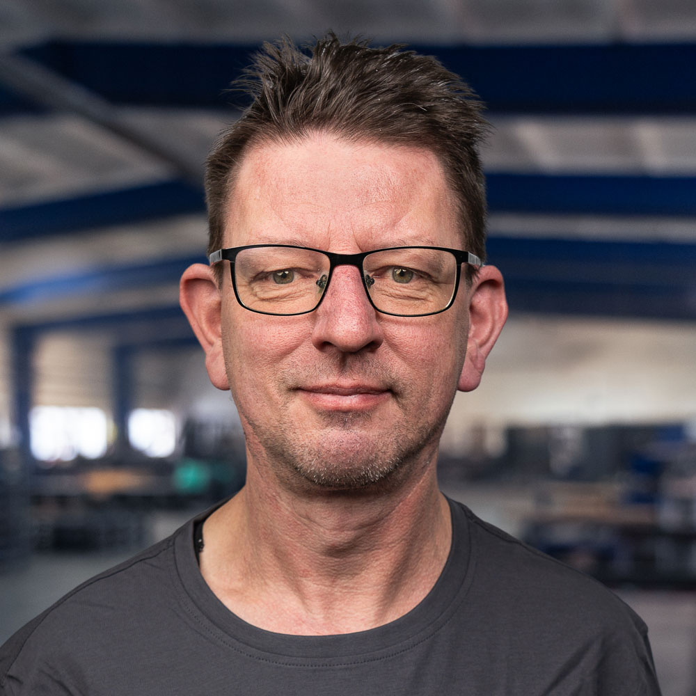 Ein Mann mittleren Alters mit kurzen braunen Haaren, Brille und einem leichten Lächeln steht in einem Raum und trägt ein dunkles T-Shirt. Der Hintergrund ist unscharf und scheint eine Industrie- oder Lagerhalle zu sein.